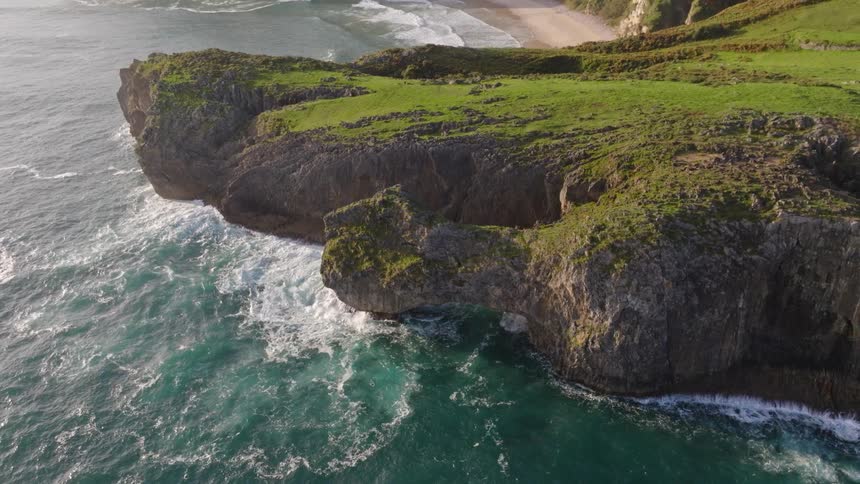 An aerial top-down view of a rocky cliff pulls back to reveal a stunning coastline with a natural rock arch, secluded beach, and distant mountains