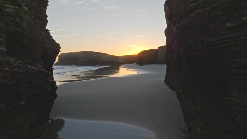 A tranquil sunset over a beautiful sandy beach, revealed by a slow pullback shot between stunning rock formations