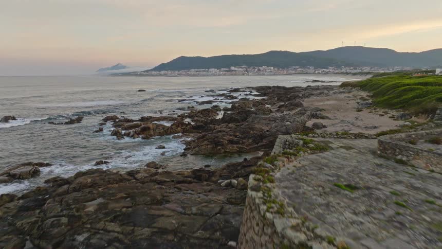 Aerial view slowly pushes forward over a historic stone fort on a rocky coast as waves crash, revealing a scenic town in the distance at dusk