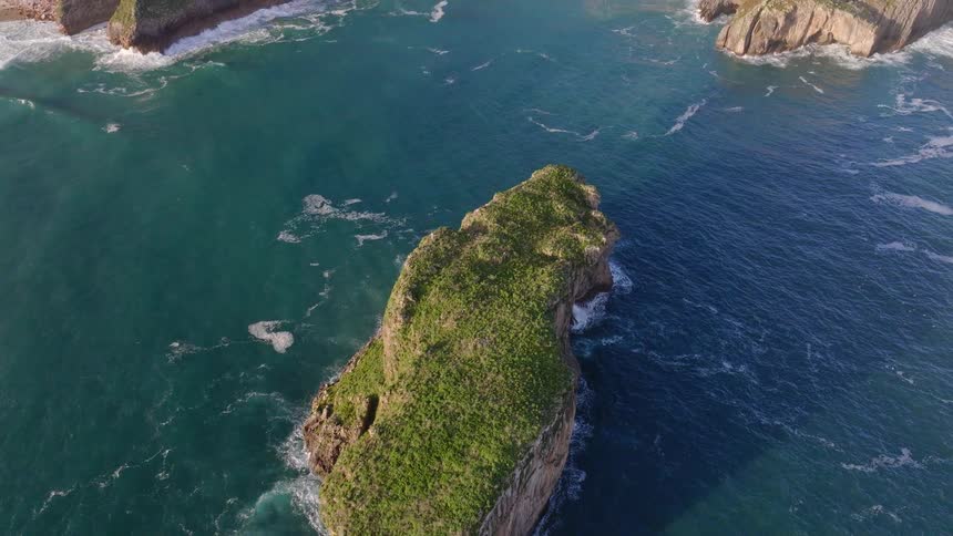 An awe-inspiring aerial view starts above a lone sea stack and tilts up to reveal a beautiful hidden cove with a sandy beach and rolling green hills