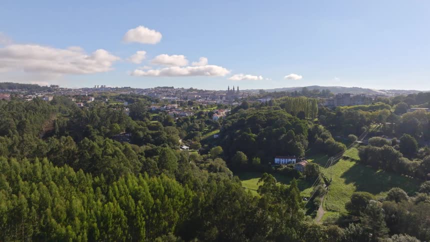 Cinematic aerial reveal of the city of Santiago de Compostela, Spain, rising over a lush green forest on a sunny day