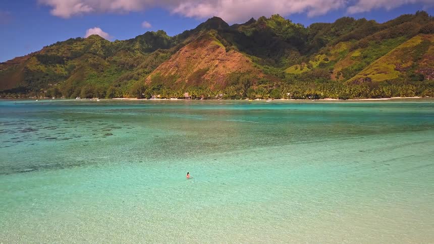A woman wades in the crystal-clear turquoise lagoon of a lush tropical island with majestic mountains