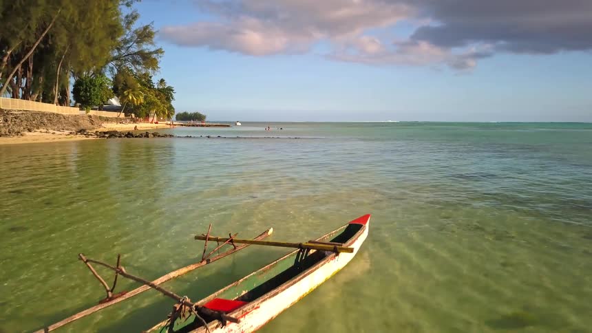 An aerial view pulls back to reveal a traditional outrigger canoe floating in the calm, clear turquoise waters of a tropical island