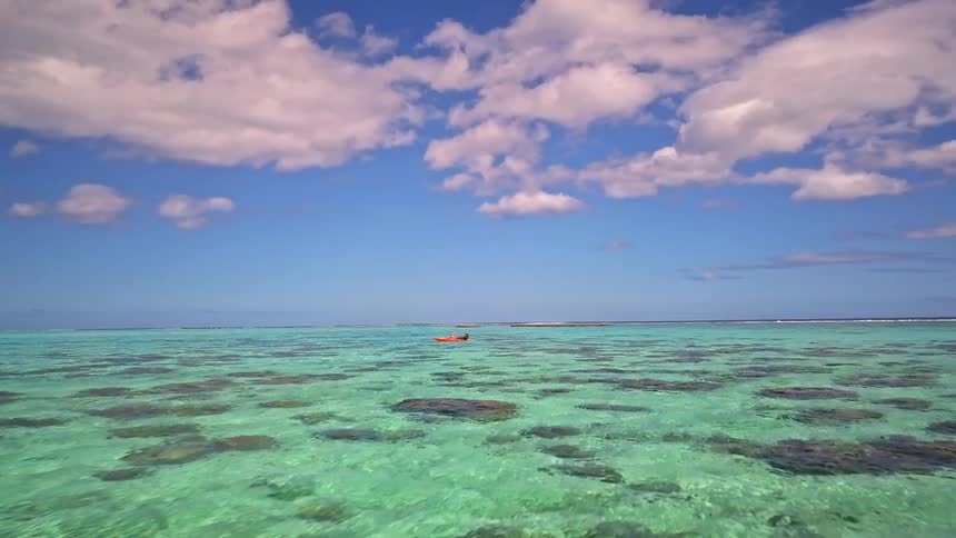 An aerial view flies slowly over the rocky shore of a tropical island towards a lone kayaker in the calm, turquoise ocean