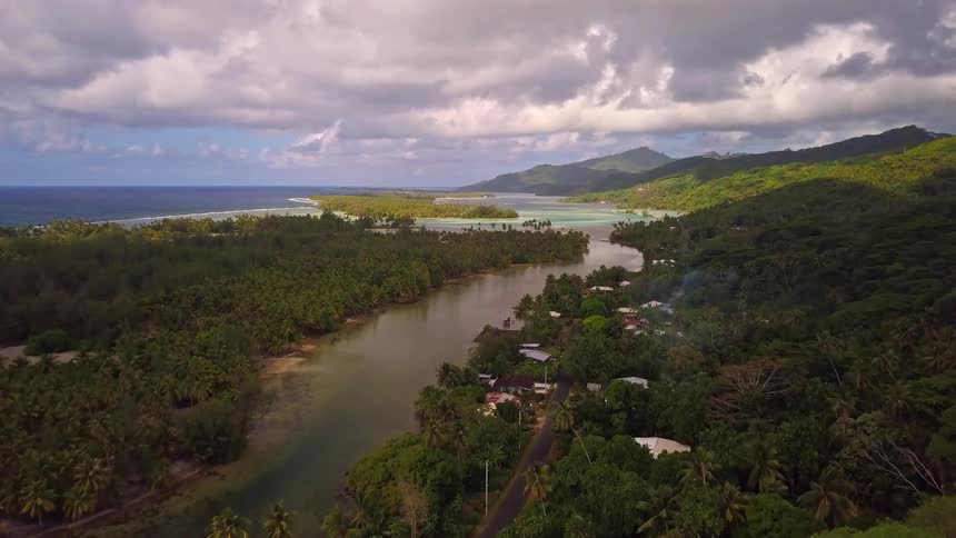 A stunning aerial view slowly pushes over a lush green jungle to reveal a winding river, a coastal village, and the vast blue ocean on a tropical island