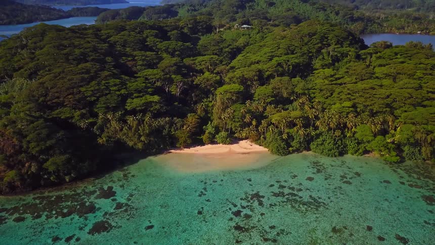 An aerial shot pulls back from a secret sandy cove with turquoise water to reveal a stunning tropical island paradise covered in lush forest