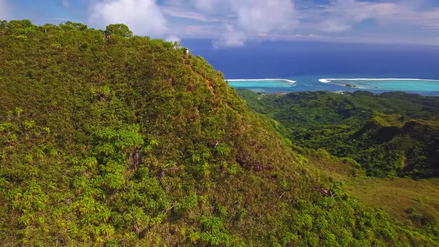 An aerial shot pulls back from a lone hiker on a lush green mountain ridge, revealing a stunning tropical island and turquoise lagoon below