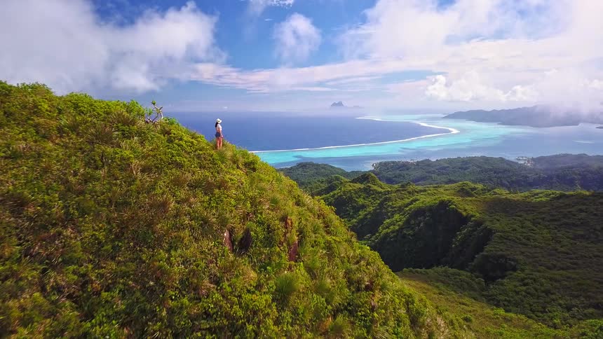 A slow aerial pullback reveals a female hiker on a mountaintop enjoying the stunning panoramic view of a tropical island paradise