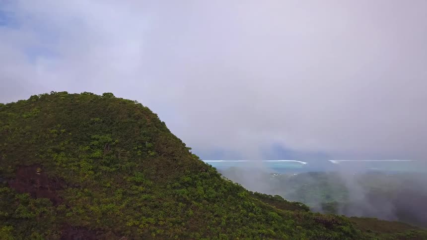 Aerial drone pulls back from a lush green mountain peak to reveal a beautiful tropical valley and ocean shrouded in misty clouds