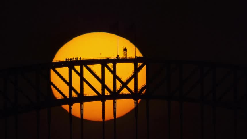 A stunning time-lapse shows the huge, glowing orange sun setting behind the silhouetted Sydney Harbour Bridge