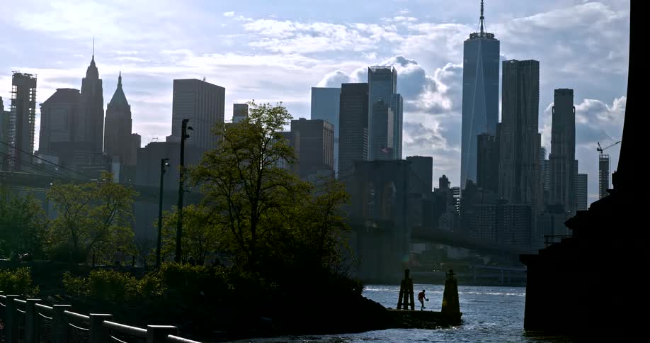 A father and child share a playful moment on a pier, silhouetted against the iconic New York City skyline with the Brooklyn Bridge in the background
