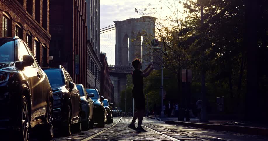 A young man is silhouetted by the setting sun as he juggles a soccer ball on a cobblestone street in Dumbo, with the iconic Brooklyn Bridge in the background
