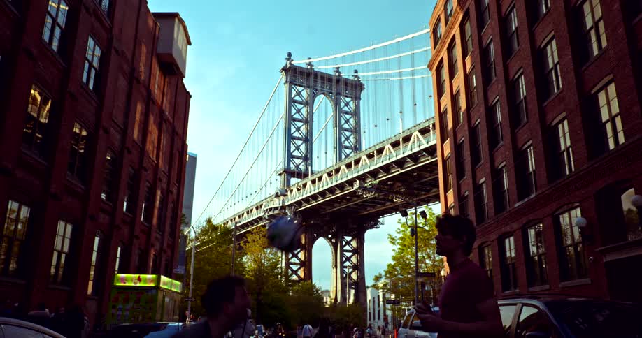 Two men juggle a soccer ball in slow motion on a street in Dumbo, Brooklyn, with the iconic Manhattan Bridge framed in the background