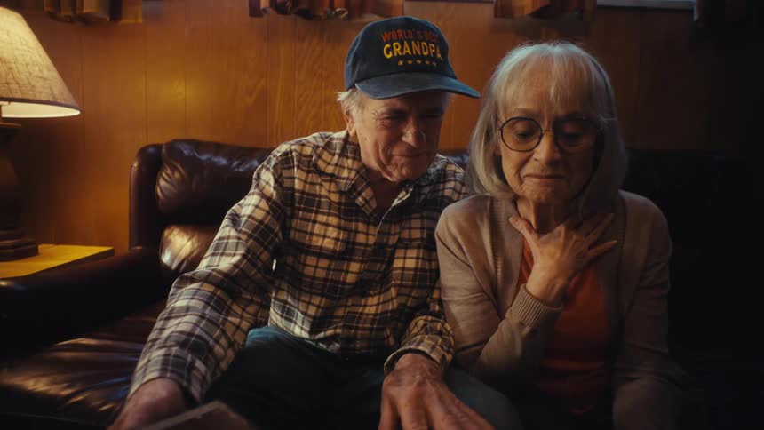 An elderly couple gets emotional as they sit together on a couch looking through an old photo album and reminiscing about past memories