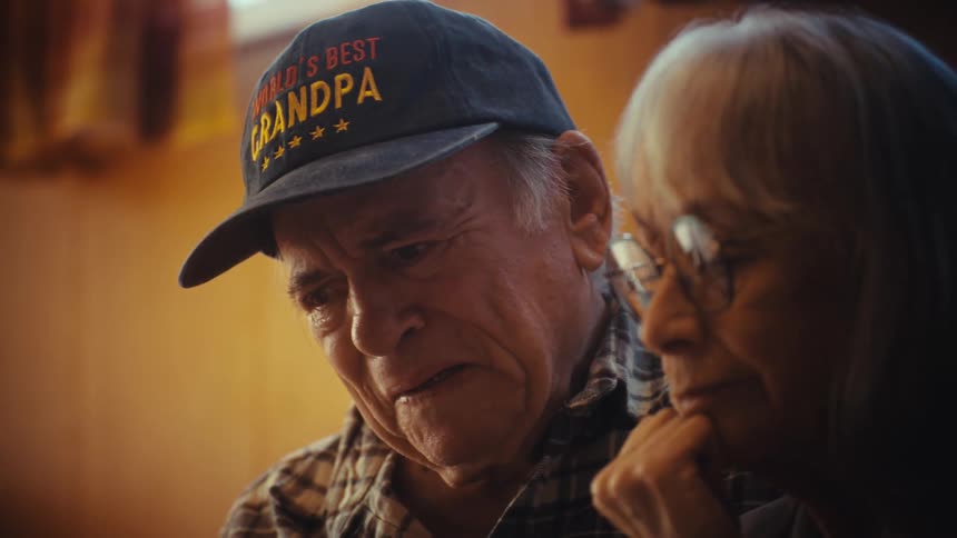 Close up of a pensive elderly couple, the man wearing a 'World's Best Grandpa' hat, sharing a quiet, sentimental moment