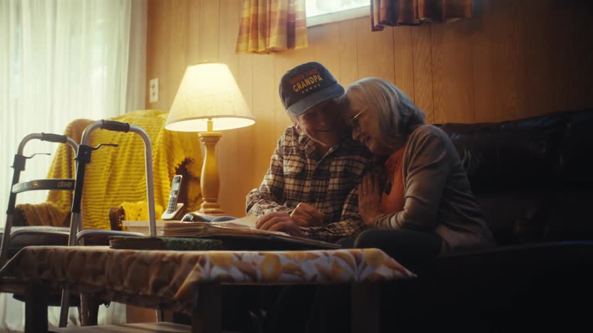 A loving elderly couple reminisces over old memories while looking through a photo album together on their living room couch