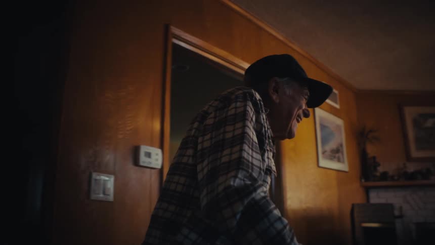 A smiling elderly man with a walker slowly emerges from a dark hallway into a warm, sunlit living room to join his wife