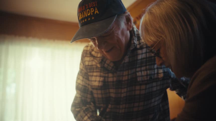 A sweet elderly couple shares a loving moment as the wife helps her husband with a walker sit down in their sunlit living room