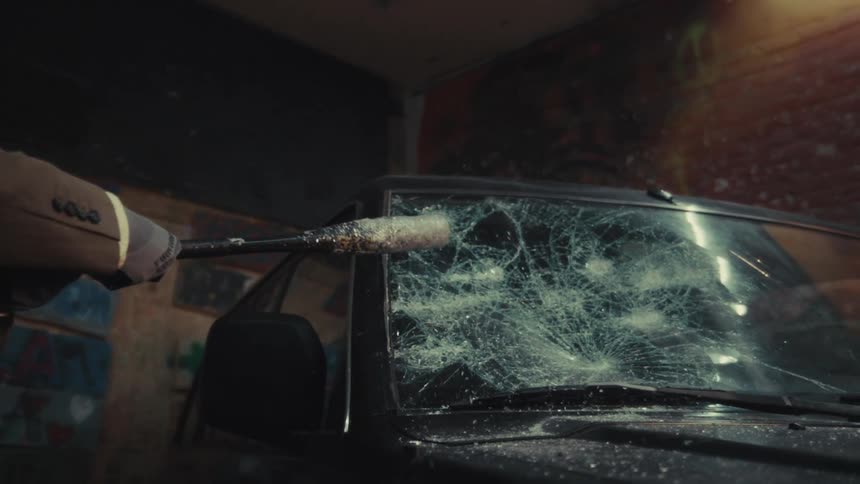 A man in a suit and protective mask aggressively smashes a car windshield with a baseball bat in slow motion