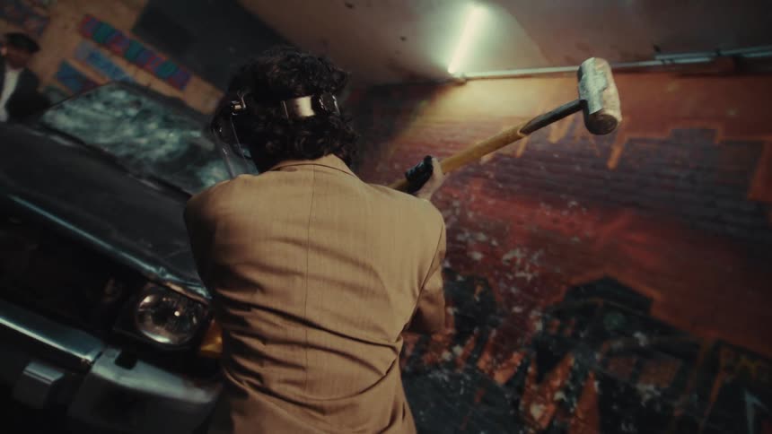 A man in a tan suit swings a sledgehammer in slow motion, preparing to smash a car in a graffiti-covered rage room