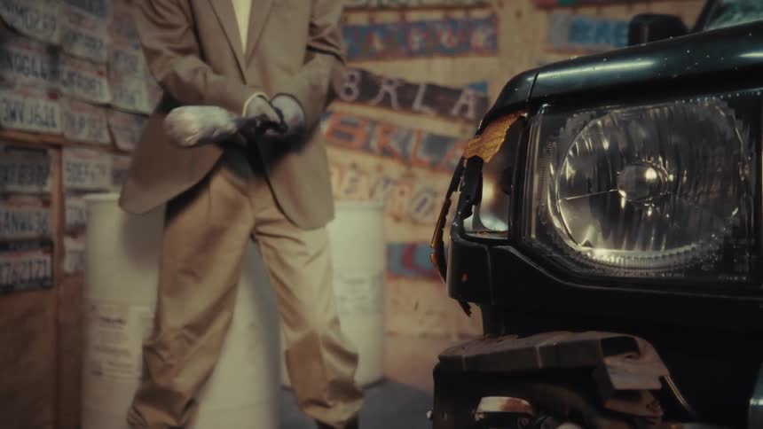 An angry man in a suit smashes a car's headlight with a baseball bat in a rage room, filmed in slow motion