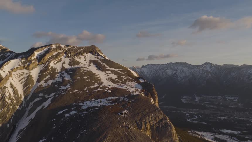 A stunning aerial shot pulls back from a rugged, snow-dusted mountain peak during a golden hour sunset
