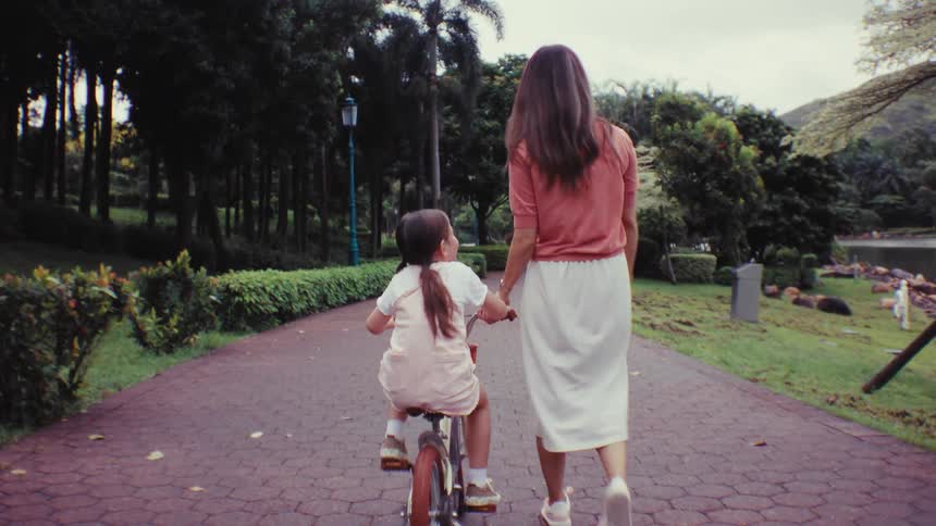 A mother holds her daughter's hand and helps her ride a bike for the first time on a path through a lush green park