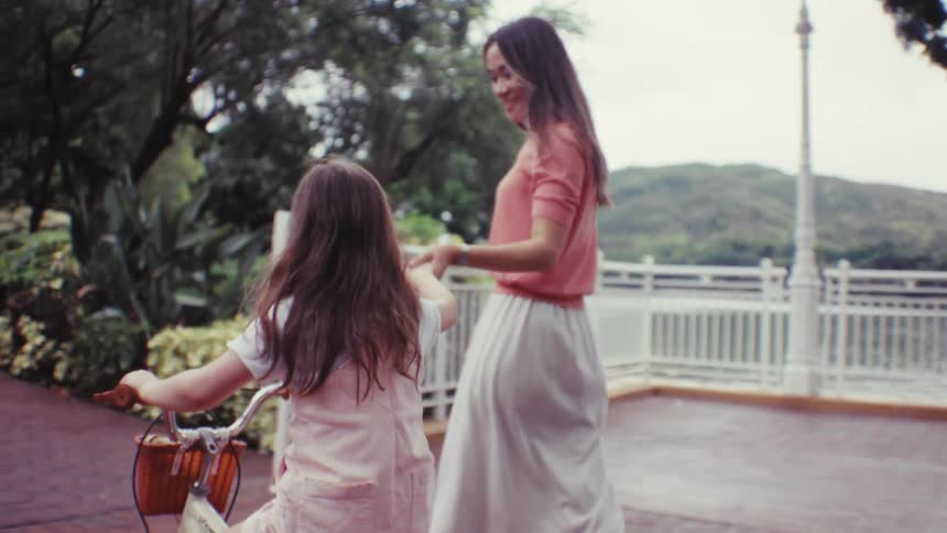 A joyful young girl riding her bicycle on a park path looks back and holds hands with her mother walking alongside her