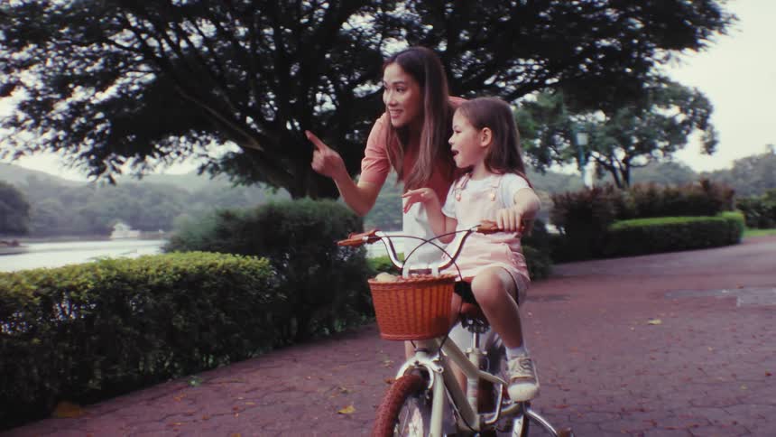 A mother and her young daughter pause on a park path while bike riding to point at something interesting by the lake