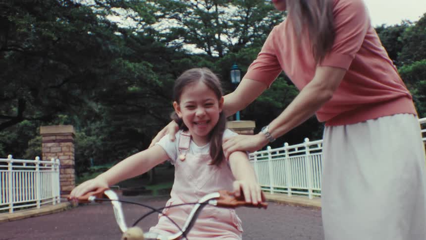 A loving mother encourages her happy daughter learning to ride her first bicycle in a beautiful park, sharing a kiss in slow motion