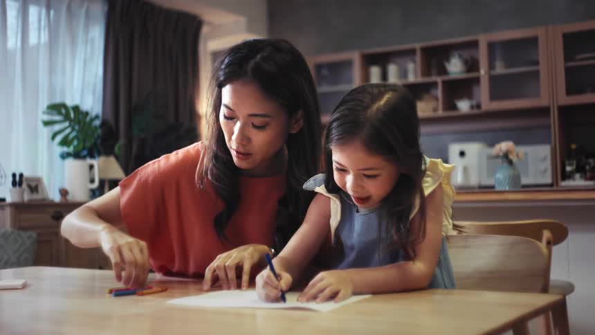 A caring mother watches and guides her happy young daughter as she draws with crayons at the dining room table in their home