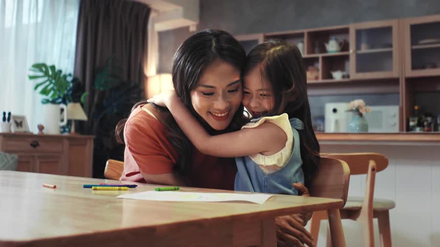 A young daughter gives her loving mother a big, warm hug at the dining table in their home in a heartwarming slow motion clip
