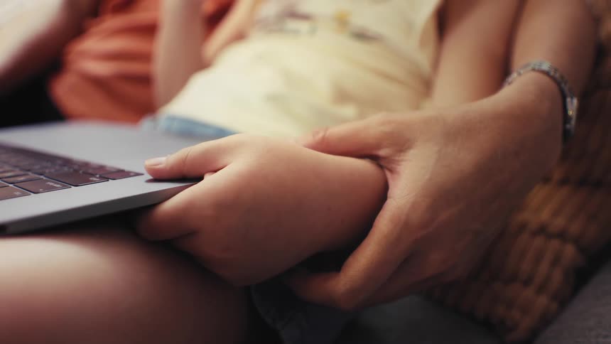 A tender close-up shows a parent's hand lovingly holding their child's hand while they look at a laptop together
