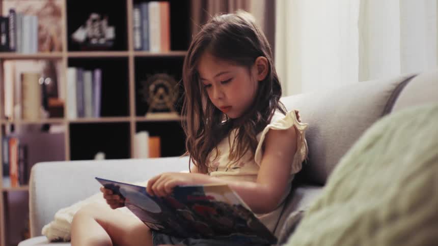 A young girl with long hair sits on a comfy couch, completely absorbed in reading a colorful picture book in her living room