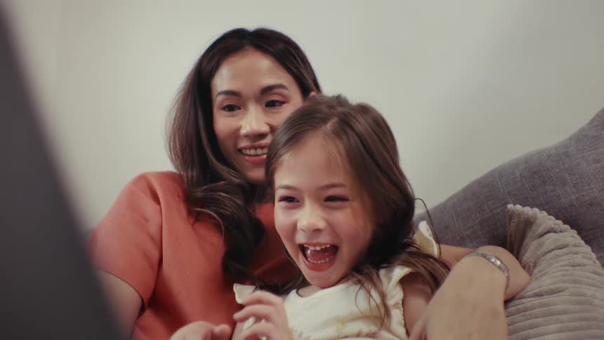 A close-up slow-motion shot of a joyful little girl laughing hysterically while watching a movie with her mother on the couch