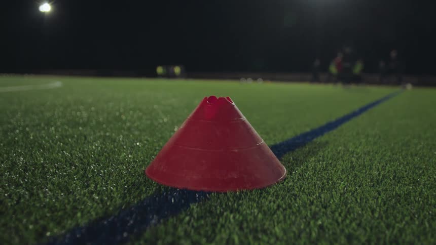 A close-up of a red training cone on an artificial turf soccer field at night with players practicing in the background