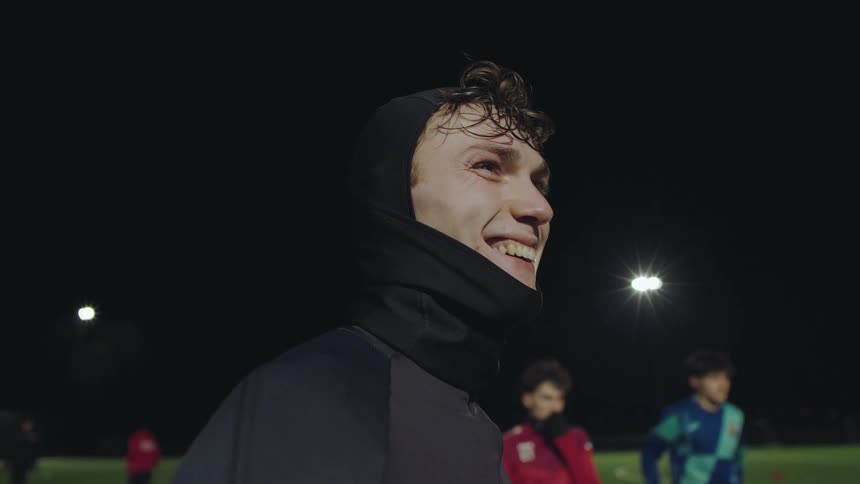 A focused young soccer player in a black balaclava breaks into a wide smile on the field during a nighttime game