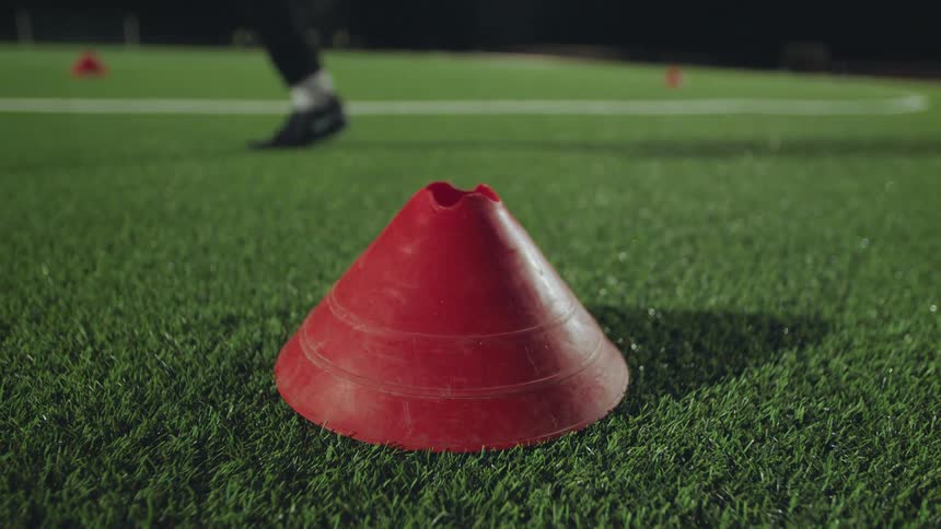 A red training cone sits on a green turf field at night as an athlete runs drills in the background