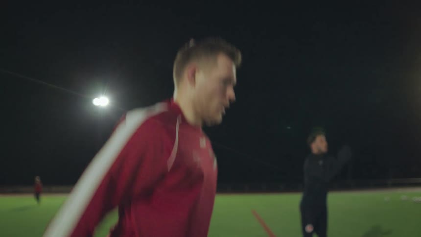 Male athlete in a red tracksuit warms up with squats and jumps on a floodlit sports field at night in slow motion