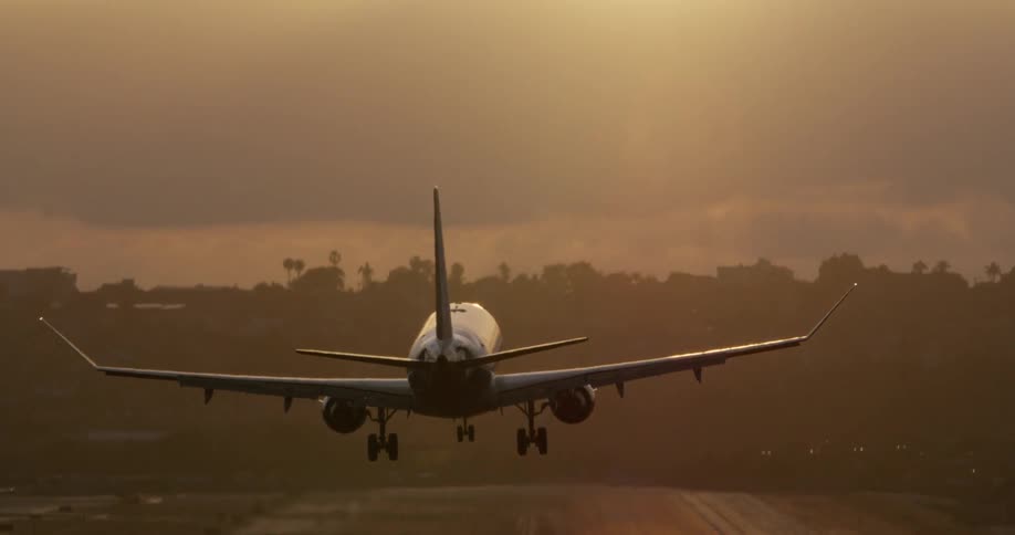 A commercial passenger jet is silhouetted against the setting sun as it descends in slow motion to land on a runway