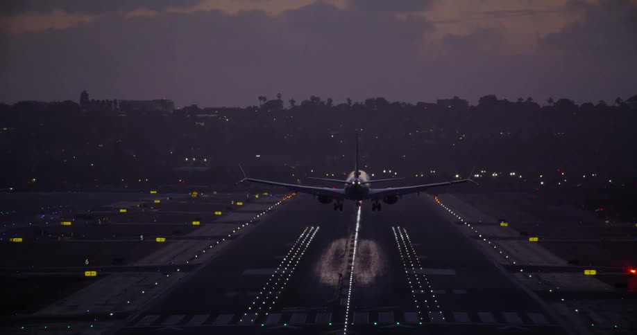 A passenger jet makes its final approach and touches down on an airport runway illuminated by bright lights at dusk