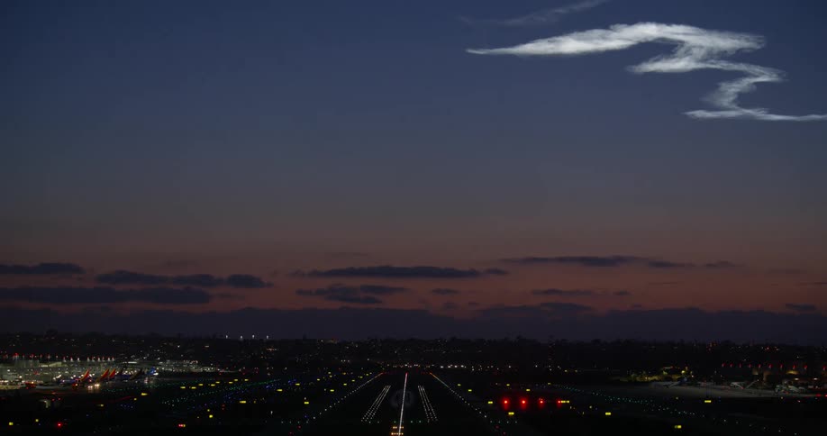 A wide shot of an active airport runway at dusk with flashing lights and the illuminated exhaust trail from a rocket launch in the sky