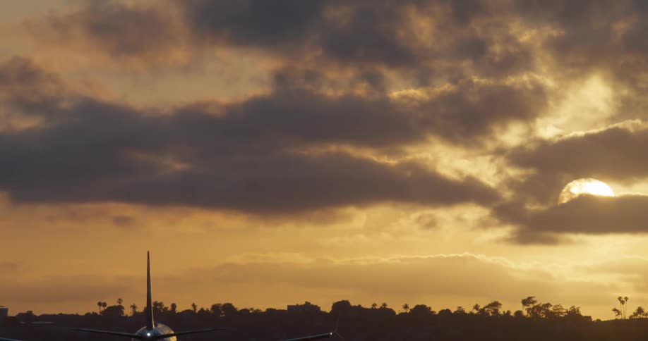 A passenger airplane descends for landing in slow motion against a dramatic golden sunset and dark clouds