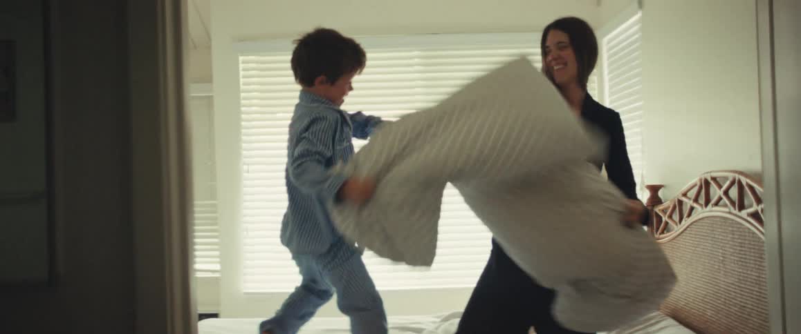 A happy mother and her son enjoy a playful pillow fight in slow motion on a bed in a sunlit bedroom