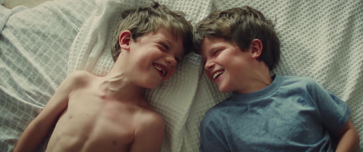 Two smiling boys lie face to face on a bed sharing a playful, intimate moment indoors