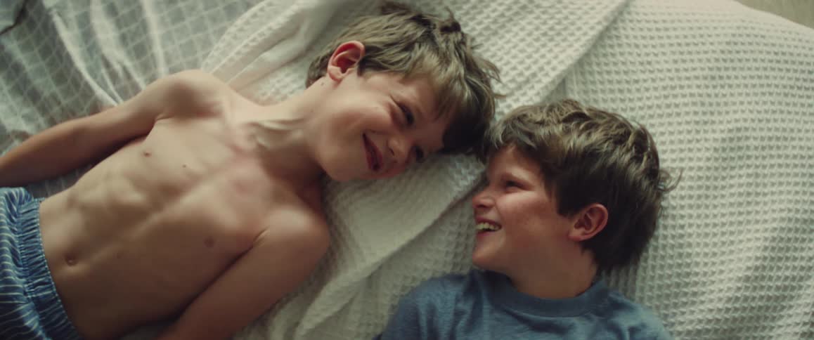 Two smiling boys lie on a white bed sharing a playful affectionate moment at home