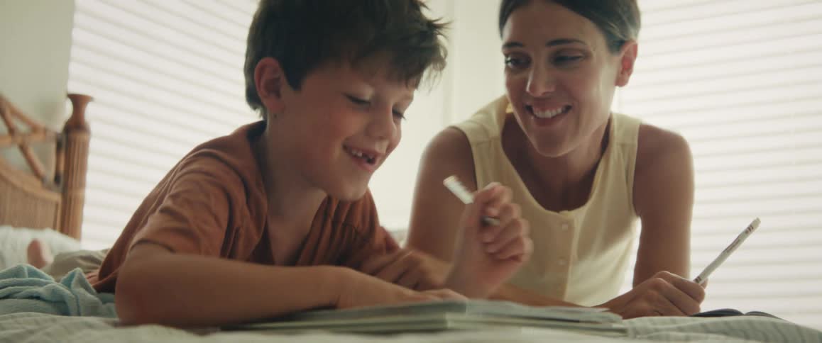 A loving mother helps her smiling son with his homework as they lie together on a bed in a sunlit room