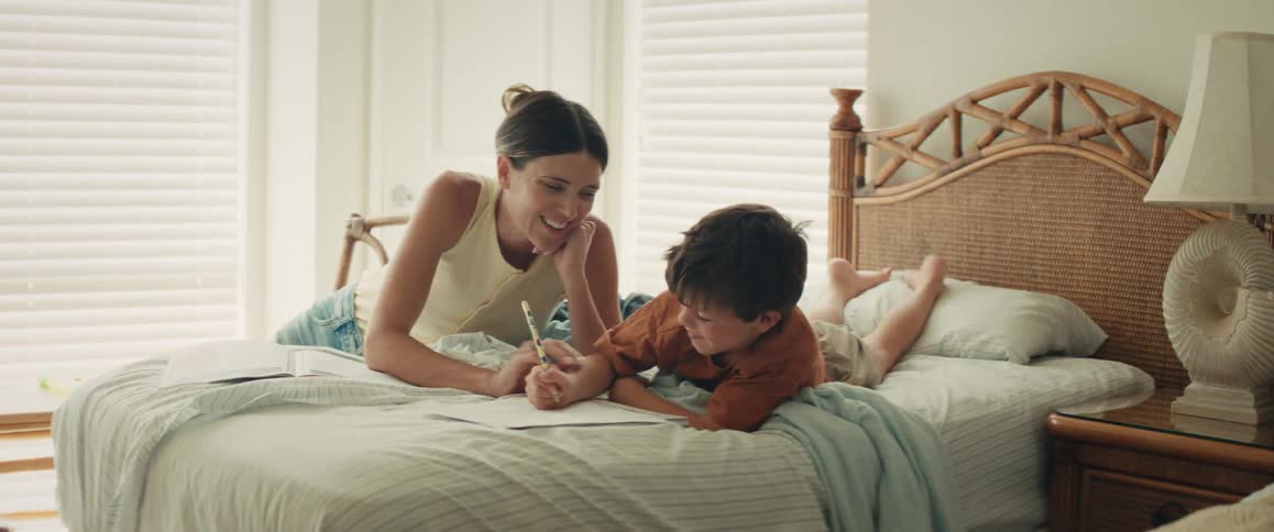A loving mother smiles as she helps her young son draw while they relax on a bed together in a sunlit bedroom
