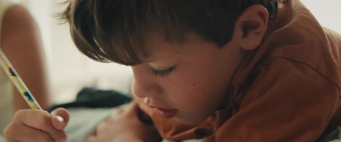 A young boy with brown hair intensely concentrates while drawing with a pencil in a close-up shot