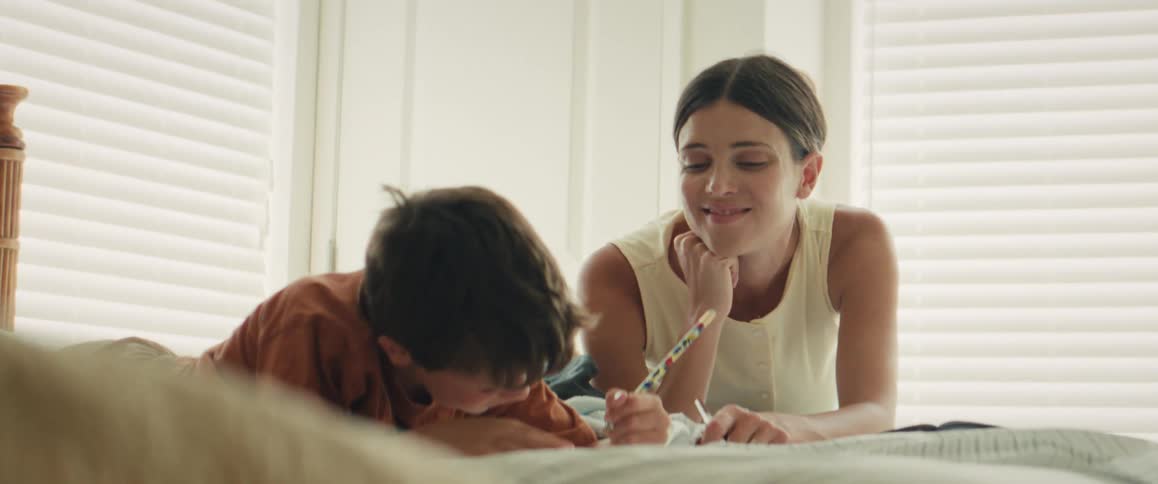 A loving mother and her young son share a happy moment drawing and laughing together while lying on a bed in a bright bedroom